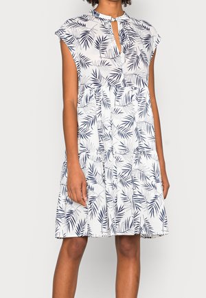 White sleeveless dress with navy blue tropical leaf print, keyhole neckline, and ruffled skirt worn by person standing against white background.