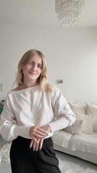 Witte off-shoulder sweatshirt met geribbelde manchetten en zwarte broek. De achtergrond bevat een witte bank met kussens en een kroonluchter erboven.