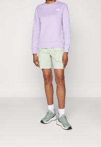 Felpa lilla con logo bianco, abbinata a pantaloni cargo verdi menta. Scarpe sportive grigio-verdi completano il look, con suole texture.