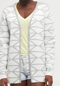 Lightweight white cardigan with a geometric gray pattern, open front design, and textured fabric. Worn over a yellow v-neck shirt and denim shorts.