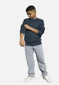 Ragazzo che arrotola la manica del suo maglione a coste blu scuro, indossando jeans azzurri e sneakers bianche, in piedi su uno sfondo bianco.