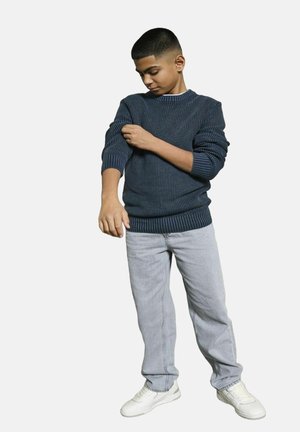 Un garçon remonte la manche de son pull côtelé bleu foncé, portant un jean bleu clair et des baskets blanches, se tenant sur un fond blanc.
