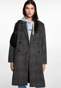 Grau karierter Übermantel mit doppelter Knopfleiste, großen notched Revers, strukturiertem Stoff und zwei Knöpfen. Über einem grauen Kapuzenpullover getragen.
