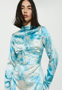 Un abito a maniche lunghe in satin azzurro chiaro con un dettaglio a fiocco in vita, caratterizzato da un motivo simile a nuvole in blu scuro e accenti beige.