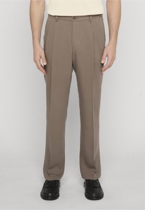 Mężczyzna w taupe plisowanych spodniach i czarnych mokasynach, stojący na tle jednolitego jasnego tła.