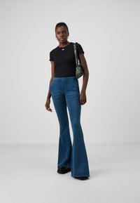 Black fitted t-shirt with a small red logo, paired with blue flared pants. The outfit is styled with a green patterned bag and black shoes.