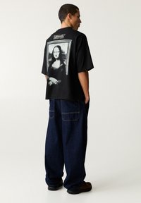 Schwarzes, übergroßes T-Shirt mit einem weißen Grafikdruck der Mona Lisa auf dem Rücken, kombiniert mit lockeren, dunkelblauen Jeans und schwarzen Schuhen.