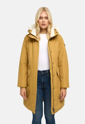 Blonde vrouw die een mosterdgele winterjas met capuchon en witte fleece voering draagt over een wit overhemd en blauwe jeans.