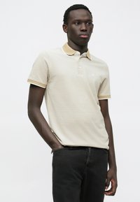 Beige polo skjorte med gul krage og ermeakenter, kortermet, og en subtil tekstur. Brukes med sorte jeans.