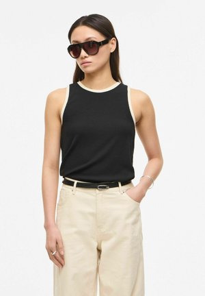 Žena u crnom topu bez rukava s bež obrubom, bež hlačama, crnim remenom, velikim crnim sunčanim naočalama i srebrnim nakitom, pozira s jednom rukom u džepu.