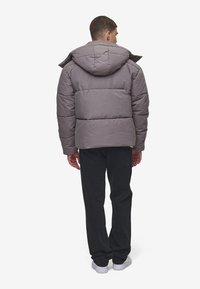 Chaqueta acolchada gris con capucha, con un diseño acolchado. Se lleva con pantalones negros y zapatos blancos, visto desde atrás.