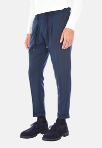 Pantaloni tailored blu navy con fibbia e chiusura con cerniera, caratterizzati da due pieghe frontali. Abbinati a scarpe in suede blu navy e calze scure.