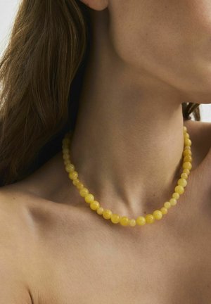 Nahaufnahme einer Person, die eine runde gelbe Perlenkette am Hals und Schlüsselbein trägt, mit sichtbarem langem braunem Haar.