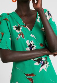 Vestido envolvente verde con estampado floral, que presenta mangas cortas abullonadas, escote en V y una textura suave con patrones florales multicolores.