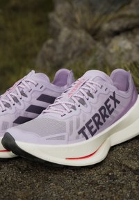Ζευγάρι ανοιχτόμωβ αθλητικά παπούτσια Adidas Terrex σε βραχώδες έδαφος με λευκές ενδιάμεσες σόλες και μαύρες εξωτερικές σόλες.