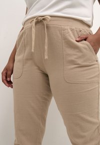 Beige jogger kalhoty vyrobené z měkké tkaniny, se splývavým pasem a zpevněným stahováním, s dvěma postranními kapsami. Texturovaný povrch.