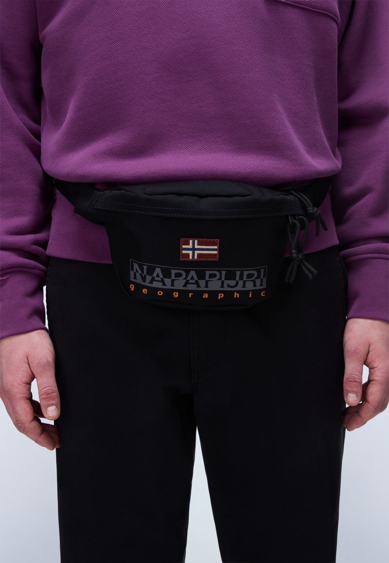 Personne portant une sacoche de taille noire Napapijri Geographic avec un patch drapeau norvégien sur un pantalon noir et une chemise violette à manches longues.