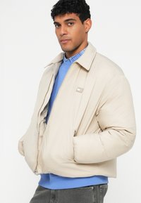 Bežinė pūkinė striukė su dideliu apykakle ir užtrauktuku. Išsiskiria minkšta medžiaga ir šoninėmis kišenėmis, dėvima virš mėlyno džemperio.