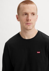Black long-sleeve t-shirt made of cotton. Features a small red Levi's logo patch on the left chest area. Round neckline, minimal design.