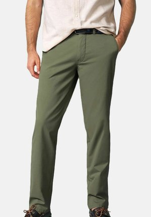 Man draagt olijfgroene broek, beige overhemd ingestopt, zwarte gevlochten riem en zwarte schoenen met bruine veters, staand met handen in de zakken.