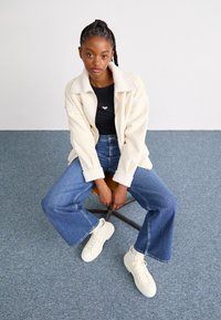 Veste en fausse laine de mouton crème sur un haut noir, associée à un jean bleu évasé et des baskets blanches. L'arrière-plan présente une texture bleue.