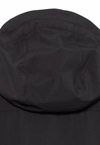 Sort stofhætte med synlige sømme og lette rynker, sandsynligvis en del af en jakke eller frakke, sat mod en ensfarvet baggrund.