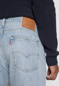 Light blue Levi's jeans with brown leather brand patch and red label, worn with a dark blue knitted sweater and a visible right hand.