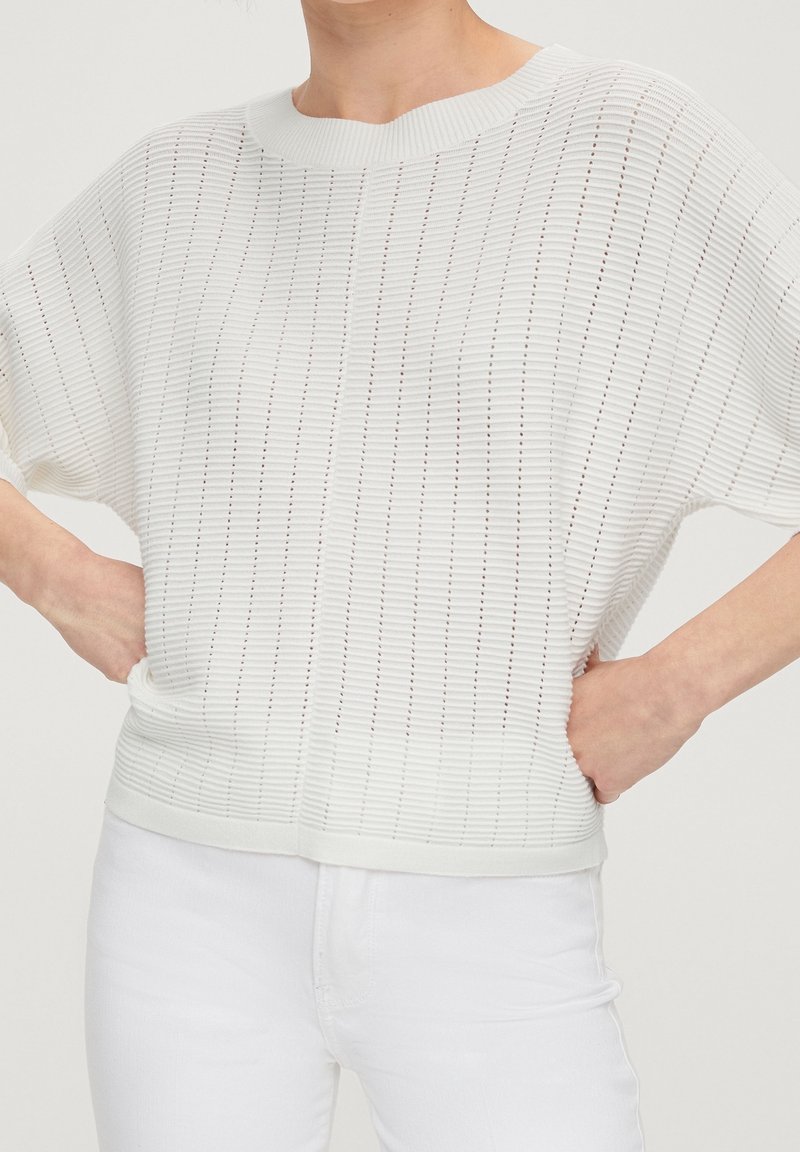 Haut tricoté blanc à coupe ample, présentant un motif texturé de rayures verticales et des manches courtes. Porté avec un jean blanc.