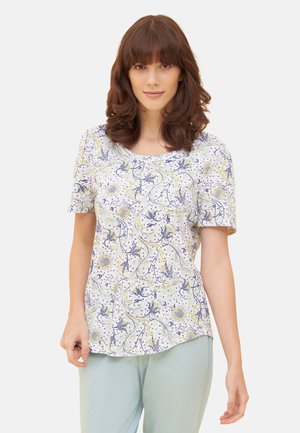 Frau mit braunen, welligen Haaren, die ein weißes Kurzarmshirt mit blauen und grünen Blumenmustern und hellblaue Hosen trägt.