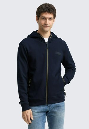 Navy zip-up hoodie with a textured fabric, gold zipper, front pocket zippers, and a fitted design, worn over a white shirt.