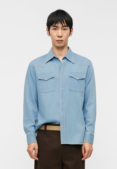 Hombre vistiendo una camisa de mezclilla azul claro con botones y dos bolsillos en el pecho, y pantalones marrón oscuro, sobre un fondo blanco liso.