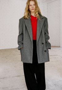 Cappotto oversized grigio in lana con colletto a revers, doppi bottoni e tasche laterali, indossato sopra un top rosso e pantaloni neri a gamba larga. Sfondo bianco.