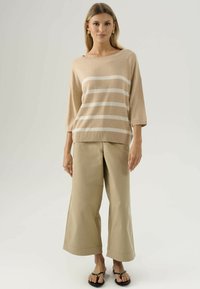 Maglione a maglia beige con strisce bianche, scollo ampio e maniche a tre quarti, abbinato a pantaloni beige a gamba larga e sandali neri.