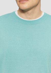 Jasnoniebieski sweter z bawełny z prążkowanym okrągłym dekoltem i teksturowanym materiałem. Nałożony na białą koszulę, ukazując gładką powierzchnię.