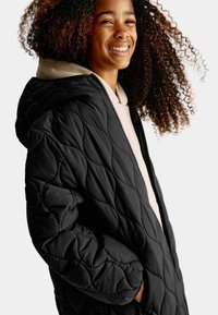 Svart vadderad jacka med huva, med ett foder inuti och framfickor med blixtlås. Slät textur med en puffig design, bärs över en ljus hoodie.