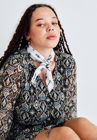 Φόρεμα με μοτίβα σε σκούρες αποχρώσεις με γεωμετρικά σχέδια, εφοδιασμένο με μακριά μανίκια και λευκή εσάρπα που δένει στον λαιμό με μαύρες λεπτομέρειες.