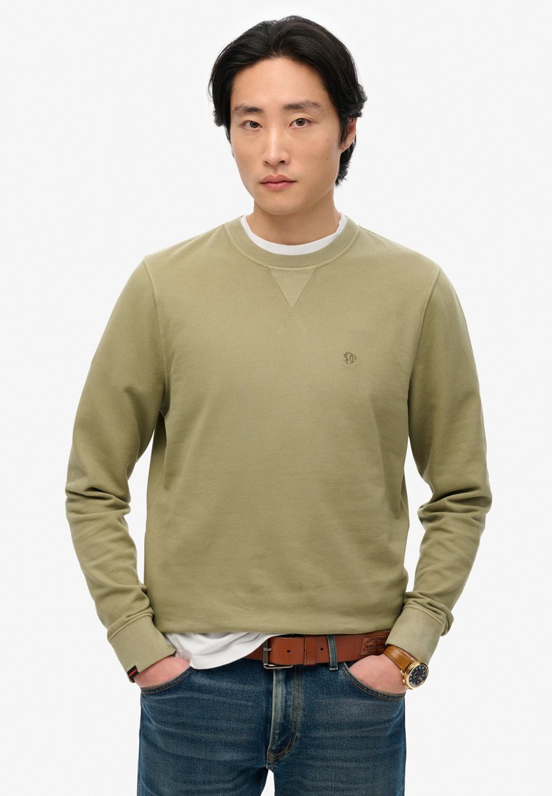 Felpa verde oliva con colletto rotondo, polsini a costine e un logo discreto sul petto, abbinata a jeans in denim azzurri chiari e una cintura marrone.