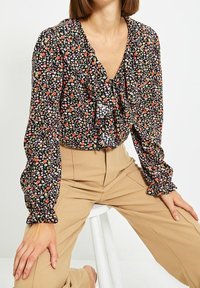 Blusa nera con fiori, con fronte arricciato e maniche a giro, caratterizzata da piccoli fiori multicolori su uno sfondo scuro, indossata con pantaloni beige.