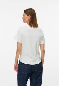 Mujer con cabello castaño que lleva pendientes de aro dorados, una camiseta blanca de manga corta sencilla y jeans oscuros, de espaldas en un fondo blanco.