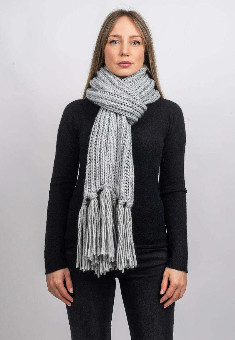 Tjock grå stickad scarf med långa fransar, har en ribbad textur. Bärs löst runt halsen och kompletterar en svart långärmad topp.