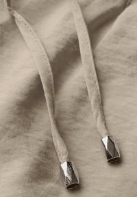 Beige stof met twee trekkoorden voorzien van metalen aglets. Het oppervlak heeft een zachte, textuurachtige uitstraling en benadrukt de stikseldetails.