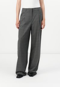 Personne portant un pantalon large à carreaux gris avec un pli avant et des chaussures noires pointues, debout devant un fond blanc.