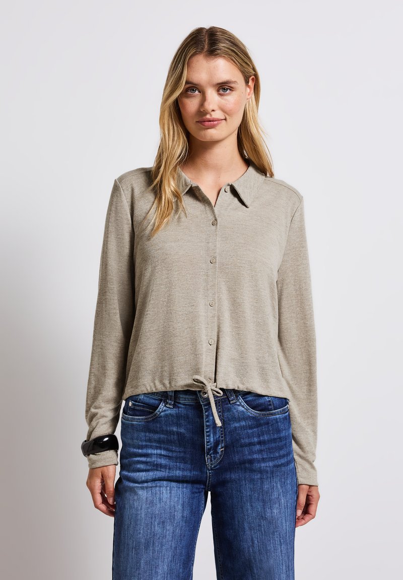 Ljusbeige långärmad topp med krage, knappstängning och dragsko i nederkant. Bärs med blåjeans. Slät textur.
