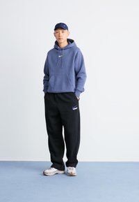 Marineblå hettegenser med snøring, sorte joggebukser med en liten logo, og hvite sneakers med brune detaljer. Modellen står mot en lys bakgrunn.