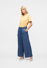 Γυναίκα που φοράει παντελόνι από τζιν με φαρδιά μπατζάκια, κίτρινο κοντομάνικο τοπ και μπεζ παπούτσια με χαμηλό τακούνι, σε απλό ανοιχτό φόντο.