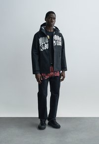 Μαύρο αδιάβροχο μπουφάν με λευκό γραφικό κείμενο, φορεμένο πάνω από μαύρο και κόκκινο πουκάμισο με σχέδιο. Συνδυασμένο με σκούρα τζιν και μαύρα αθλητικά παπούτσια.