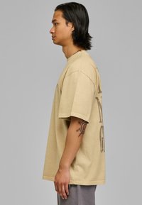 Tricou oversize bej din bumbac, cu mâneci scurte, guler rotund și un imprimeu grafic pe lateral. Textură fină.