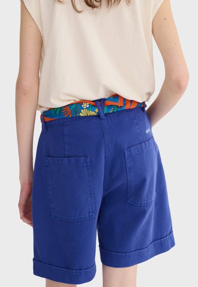 Osoba oblečená v modrých šortkách s veľkými zadnými vreckami a farebným květinovým opaskom, doplnená krémovým tielkom bez rukávov.