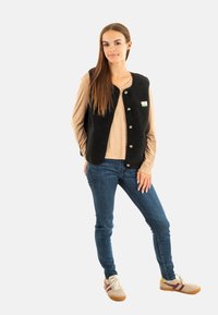 Zwarte fleece vest met knoopsluitingen, gedragen over een beige langemouwen top, gekleed met blauwe jeans en lichtgekleurde sneakers.
