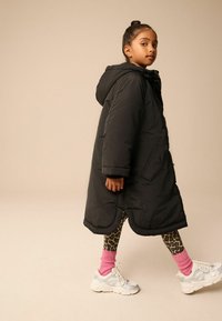 Giacca nera imbottita con cappuccio, vestibilità over, e tasche laterali; abbinata a leggings maculati, calzini rosa e sneakers argentati.